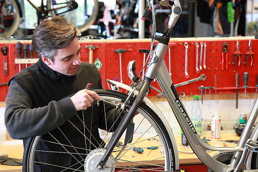 Mechatronikermangel Ein Mann beugt sich in einer Fahrradwerkstatt über das Vorderrad eines Fahrrades und zieht eine Schraube fest.