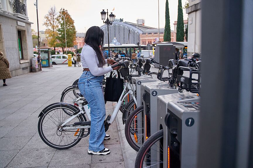 Frau entleiht Fahrrad an Bikesharing-Station Eine junge Frau mit langen dunklen Haaren, weißem Oberteil, Jeans und schwarzer Umhängetasche steht an einer Bike-Sharing-Station und entnimmt ein weißes Leihfahrrad. Die Station verfügt über mehrere nummerierte Stellplätze mit grauen Dockingelementen. Im Hintergrund sind ein belebter Stadtplatz, historische Gebäude, Passanten und ein Bahnhofsgebäude zu sehen.